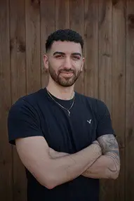 Man with arms crossed, standing against wooden fence.