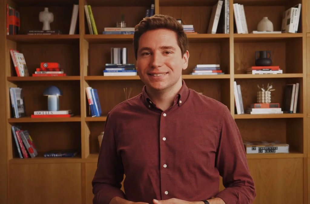 Man in maroon shirt smiles in front of bookshelf